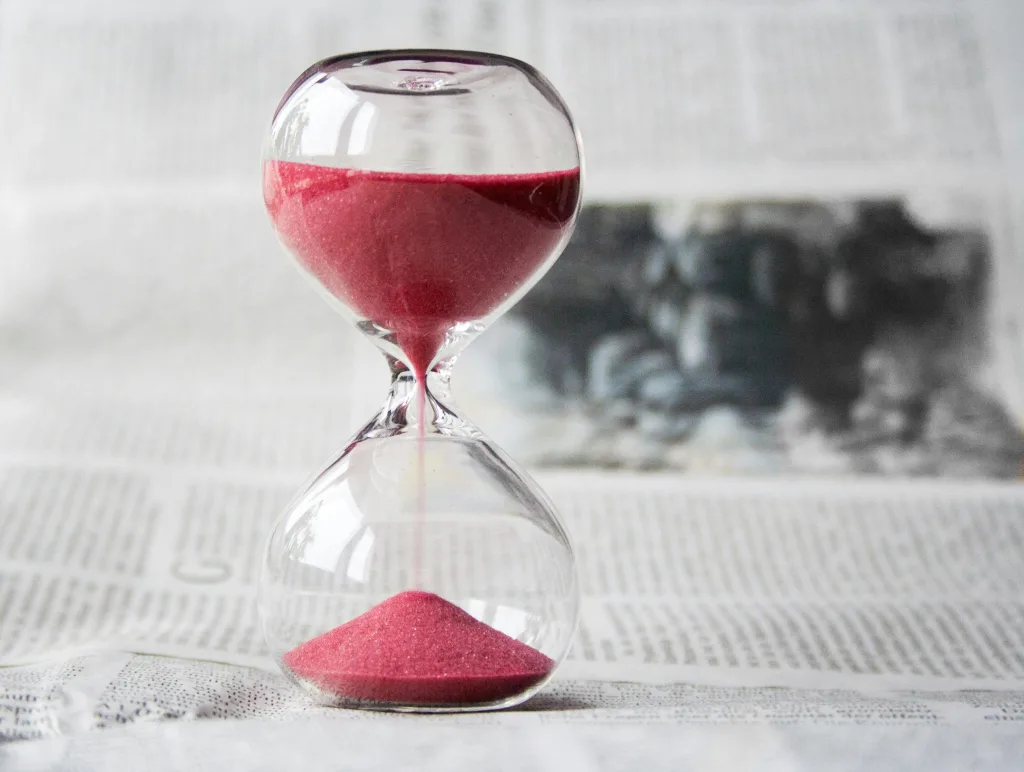 hourglass-time-hours-sand-39396-39396 Close-up of a transparent hourglass with pink sand flowing, placed on a newspaper background.